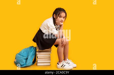 Sad Asian Schoolgirl Sitting On Books Stack Over Yellow Background Stock Photo