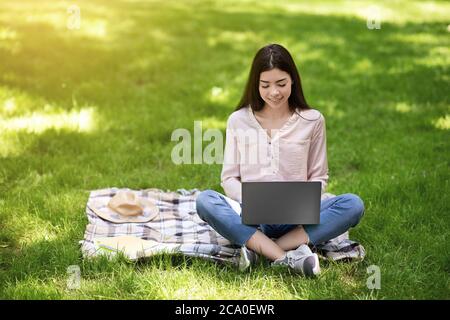 Korean Student Girl Studying Outdoors With Laptop In Park, Sitting On Lawn Stock Photo