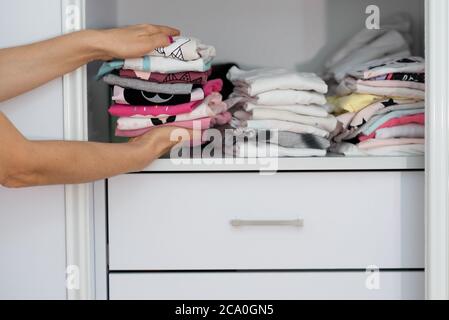 Mom picking out kid outfit at home to send her children back to school. New colored clothing stacked in a pile close-up in white wardrobe. Woman folding clothes on a shelf in a closet. Stock Photo