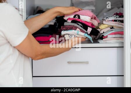 Mom picking out kid outfit at home to send her children back to school. New colored clothing stacked in a pile close-up in white wardrobe. Woman folding clothes on a shelf in a closet. Stock Photo