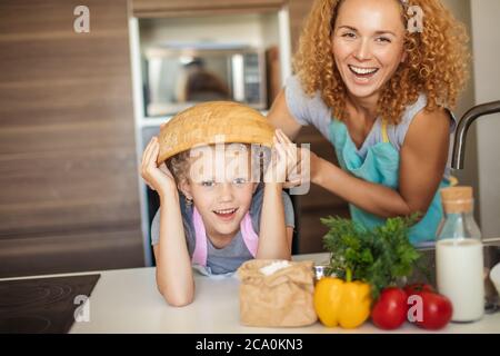 Beautiful mother and cute little daughter having fun in the kitchen while preparing to cook muffins in the bowl. Happy girl wears the bowl on head and Stock Photo