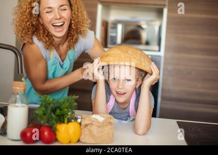 Beautiful mother and cute little daughter having fun in the kitchen while preparing to cook muffins in the bowl. Happy girl wears the bowl on head and Stock Photo