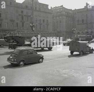 1960, historical, a Fiat 500 car and Citroen 2CV van parked on a street with an old-style fire engine, with an open-air cabin passing by, Nice, France. A  circa 1920s fire engine with its open cabin for it's driver and crew was an unusual sight for the time, perhaps why the photograph was quickly taken by an amateur by the side of the road. Stock Photo