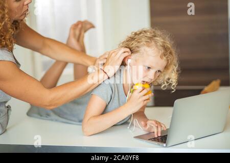 Everyday life in family with kids. Frizzy haired girl eating paprica and watching cartoons on laptop with earphones while for mother adjusting her hai Stock Photo