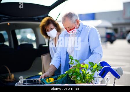 Senior couple with face masks putting shopping in car outside supermarket in city. Stock Photo