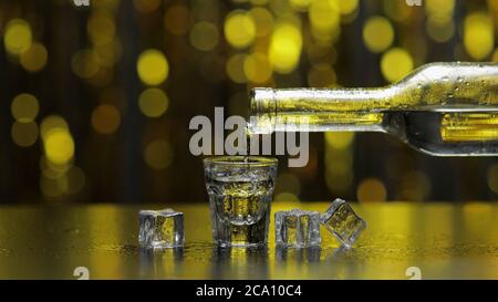 Bartender pouring up frozen vodka from bottle into shot glass with ice cubes against shiny gold party celebration background. Barman pour of cold transparent alcohol drink vodka tequila in shot-glass Stock Photo