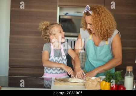 pretty little caucasian girl dressed in apron and wearing ponytail from blonde curly hair kneadling dough while cooking appetizing pie with mom, inter Stock Photo
