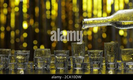 Bartender pouring up frozen vodka from bottle into eight shots glasses with ice cubes against shiny gold party celebration background. Barman pour of cold transparent alcohol drink vodka tequila Stock Photo