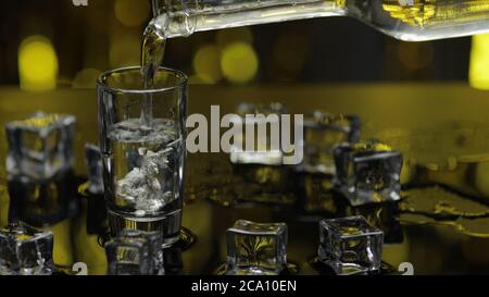 Bartender pouring up frozen vodka from bottle into shot glass with ice cubes against shiny gold party celebration background. Barman pour of cold transparent alcohol drink vodka tequila in shot-glass Stock Photo