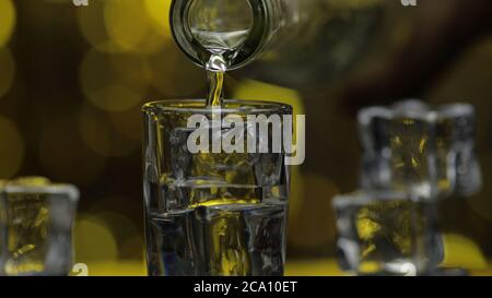 Bartender pouring up frozen vodka from bottle into shot glass against shiny gold party celebration background. Barman pour of cold transparent alcohol drink vodka tequila in shot-glass Stock Photo