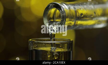 Bartender pouring up frozen vodka from bottle into shot glass against shiny gold party celebration background. Barman pour of cold transparent alcohol drink vodka tequila in shot-glass Stock Photo