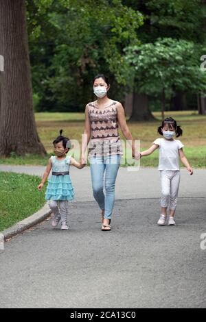 An Asian American family, likely Chinese, with 2 seriously cute daughters walk in the park holding hands and wearing surgical masks. In Queens, NYC. Stock Photo