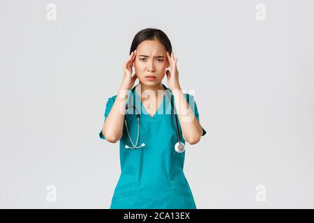 Covid-19, healthcare workers, pandemic concept. Overworked and exhausted asian female doctor, physician feeling sick, wear scrubs, touching head Stock Photo