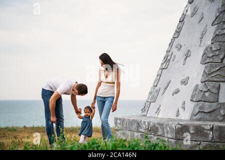 Happy young family in white t-shirts and blue jeans with a small daughter in dress near to the lighthouse Stock Photo