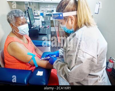 With the Corona lab tech wears mask, face shield, gloves, and disposable gown to take a blood sample from patient eho is also wearing a mask. If you think you have the virus will you go see a doctor? Stock Photo