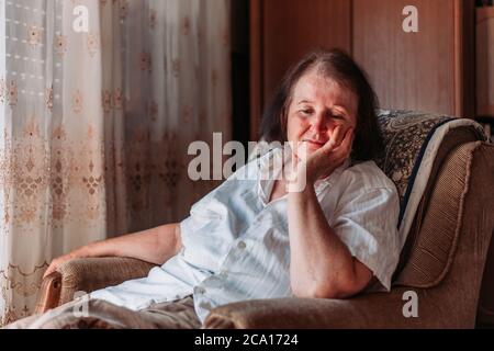 Close-up portrait of an elderly woman sitting in the chair, hand on her head, looking worried Stock Photo