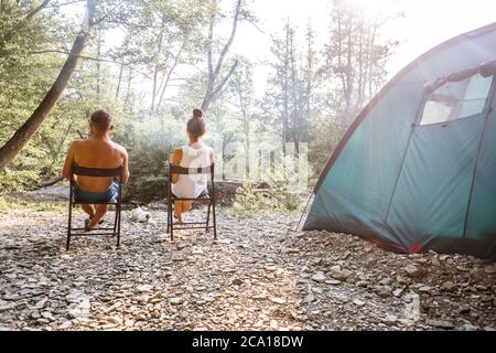 Relaxed couple sitting outside tent while camping nearby mountain river on a sunny day. Woman holding coffee talking with man. Two young caucasian people enjoying camping together. Stock Photo