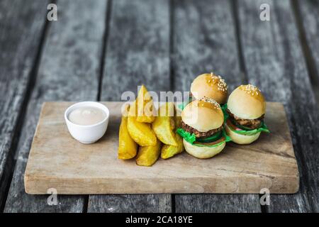 Three mini burgers with beef patty, french fries and mayonnaise on wooden board. Shabby aged table background. Unusual food serving. Fast food and unhealthy lifestyle. Stock Photo