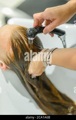 Female young red head customer wearing face mask washing hair in beauty salon by hair stylist.