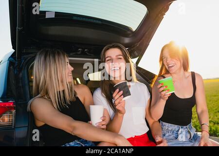 Happy road trip of three smiling women, relax at vehicle back with coffee cups take away at sunlight. Stock Photo