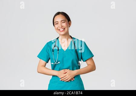 Covid-19, healthcare workers, pandemic concept. Happy pretty asian female doctor, physician in scrubs having fun, laughing and touching belly while Stock Photo