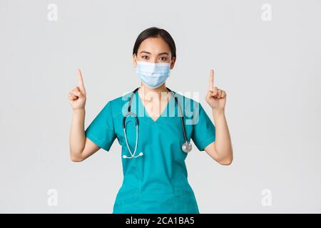 Covid-19, coronavirus disease, healthcare workers concept. Young professional asian female doctor, nurse in medical mask and scrubs, pointing fingers Stock Photo