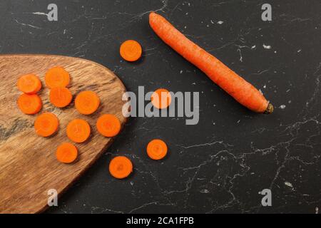 Carrot sliced to small circles and chopping board, whole one next to it on black working desk, photo from above Stock Photo