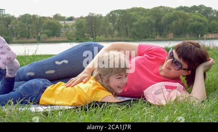 Little girl in nature, having fun eating chips. The girl has fun playing with her mother against the background of the river. Child have fun on the lake. Outdoor picnic. Stock Photo