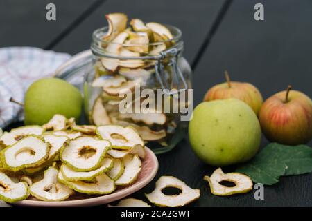 Dried apple slices in open glass jar. Homemade organic dried apple chips with fresh apple on black table background. Sweet vegan snack. Healthy and nutrition concept. Shallow depth of field Stock Photo