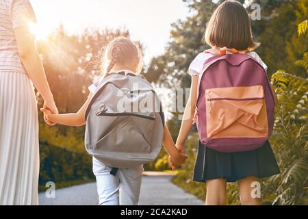 Parent and pupils of primary school go hand in hand. Woman and girls with backpacks behind backs. Beginning of lessons. First day of fall. Stock Photo