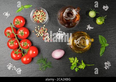 A  twig of cherry tomatoes with some coarse salt grains, an oil and a vinegar jug colorful pepper, red onion, dill, parsley and basil on a slate slab. Stock Photo