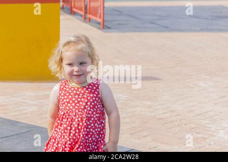 Cute little girl in red dotted dress walking on playground. One little girl walks on the playground in the street. Copy space. Summer recreation and h Stock Photo
