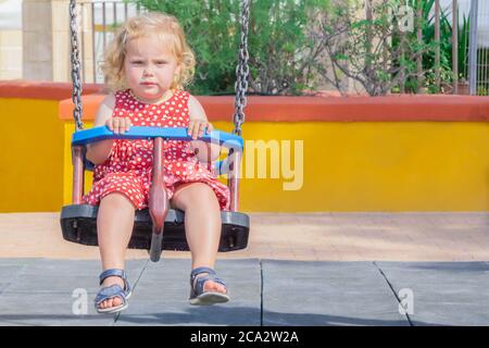 Blond little girl on swings outdoor in playground. Toddler swinging. Cute girl in red dotted dress swings in public playground. Little girl having fun Stock Photo