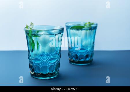 Two beautiful vintage blue glasses with cold drink and ice cubes, with fresh green mint leaves on deep blue and light blue background. Backdrop for yo Stock Photo