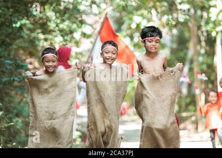 three children try to run fast and jump in the sack race with friends support them on Indonesian independence day Stock Photo