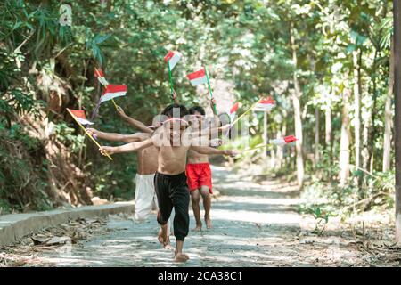 having fun group of kids running without clothes chasing each other when holding small the red and white flag and raised the flag against a tree background Stock Photo