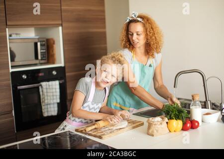 Family business. Happy mother and daughter preparing together christmas cookies flattening the dough using a rolling pin, girl is looking at camera. Stock Photo