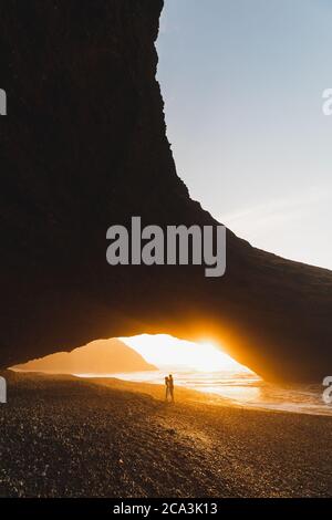 Lovely couple kissing under big arch on Legzira ocean beach and enjoying fantastic warm sunset. Famous and popular landmark in Morocco. Beautiful evening light. Stock Photo