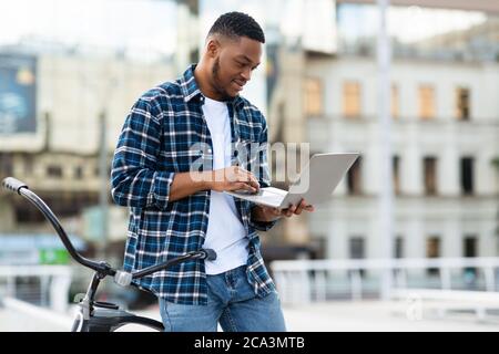 Black man using laptop, standing in downtown with bike Stock Photo
