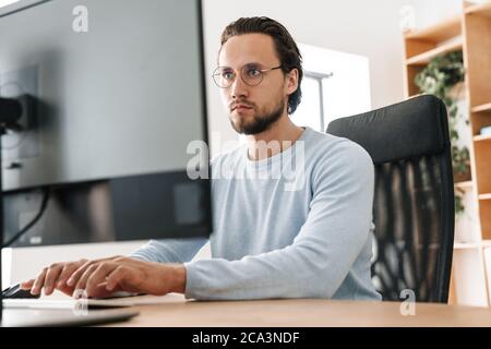 Image of focused unshaven programmer man wearing eyeglasses working with computer in office Stock Photo