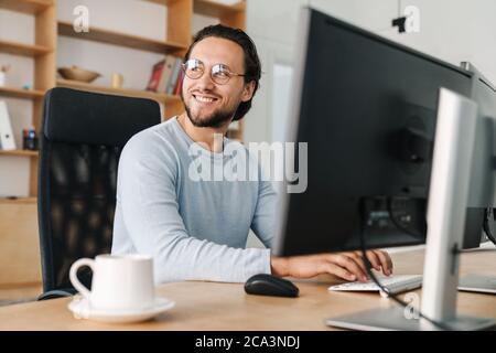 Image of smiling unshaven programmer man wearing eyeglasses working with computer in office Stock Photo