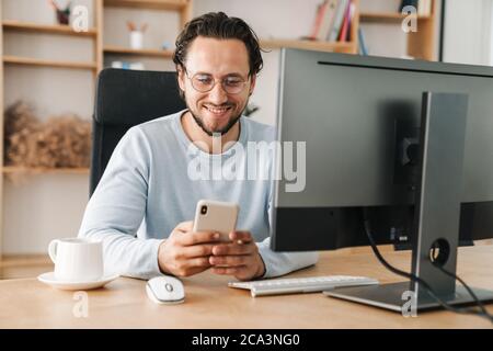 Image of smiling programmer man wearing eyeglasses using cellphone while working with computer in office Stock Photo