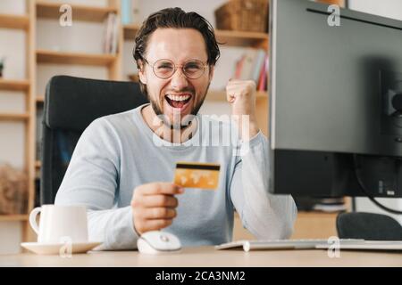 Image of excited programmer man making winner gesture and holding credit card while working with computer in office Stock Photo