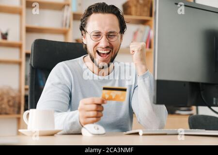 Image of excited programmer man making winner gesture and holding credit card while working with computer in office Stock Photo