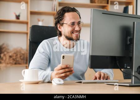 Image of smiling programmer man wearing eyeglasses using cellphone while working with computer in office Stock Photo