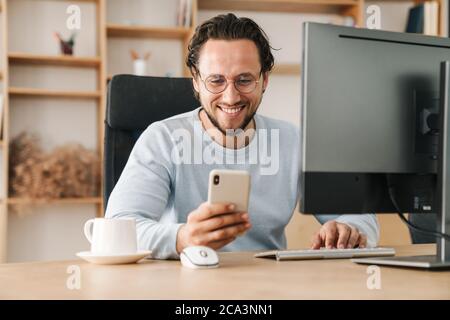 Image of smiling programmer man wearing eyeglasses using cellphone while working with computer in office Stock Photo