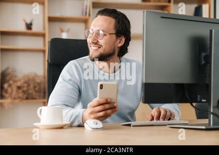 Image of smiling programmer man wearing eyeglasses using cellphone while working with computer in office Stock Photo