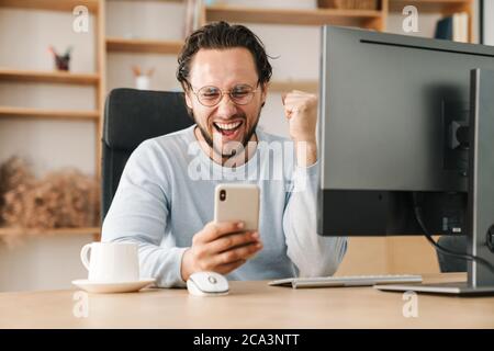 Image of excited programmer man making winner gesture and using cellphone while working in office Stock Photo