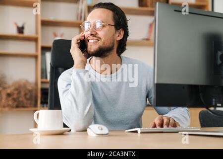 Image of smiling programmer man wearing eyeglasses talking on cellphone while working with computer in office Stock Photo
