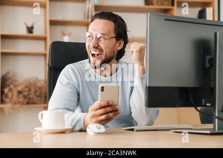 Image of excited programmer man making winner gesture and using cellphone while working in office Stock Photo